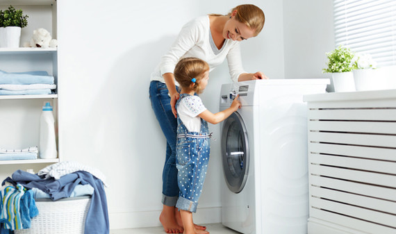 A young girl learning how to use the washing machine with guidance from her mother