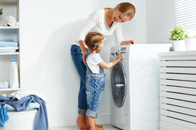 A young girl learning how to use the washing machine with guidance from her mother