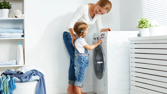 A young girl learning how to use the washing machine with guidance from her mother